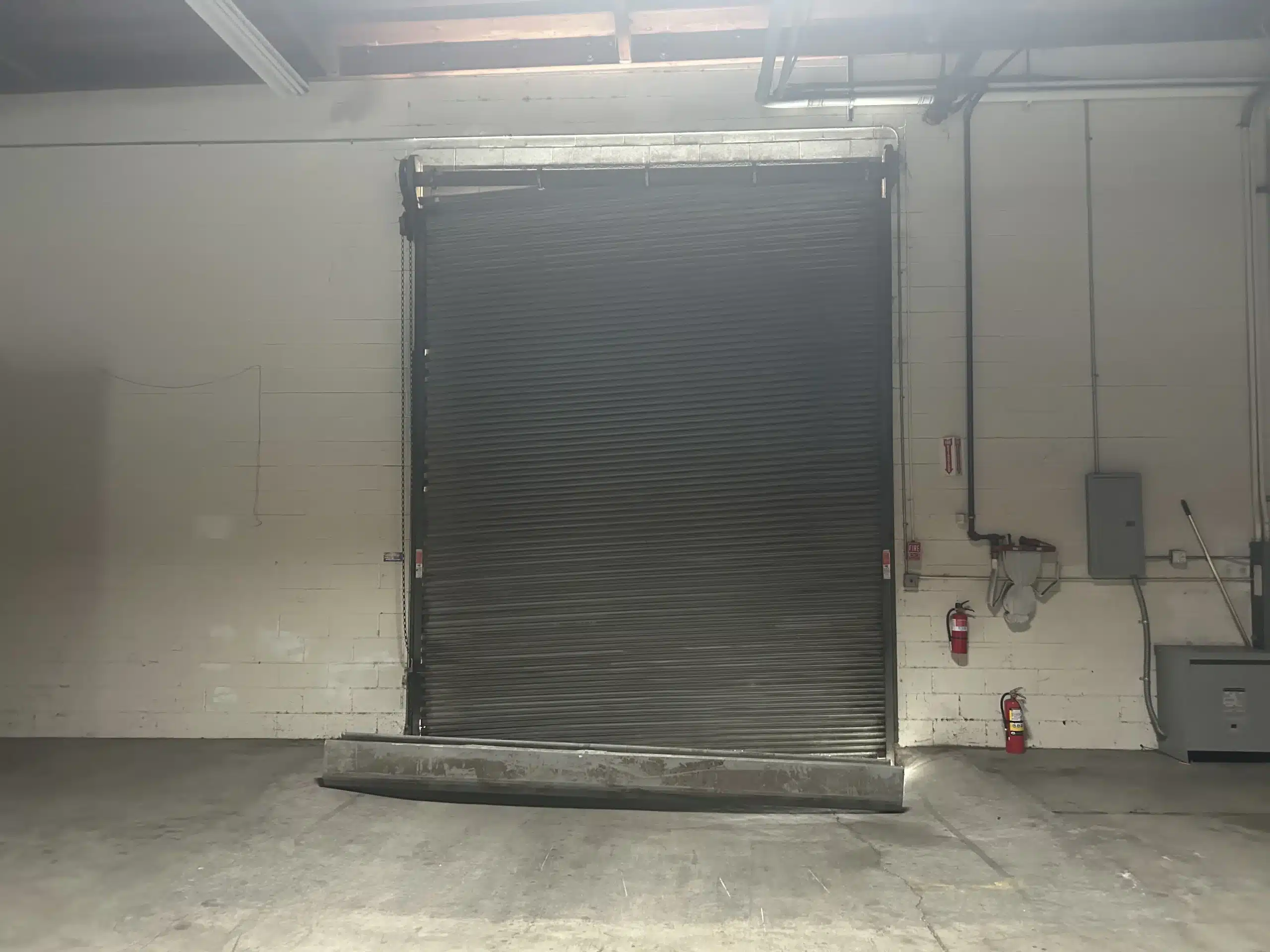 An interior view shows a heavy industrial roll-up door jammed crookedly in its opening with a dislodged bottom bar on the floor, highlighting serious commercial garage door issues.