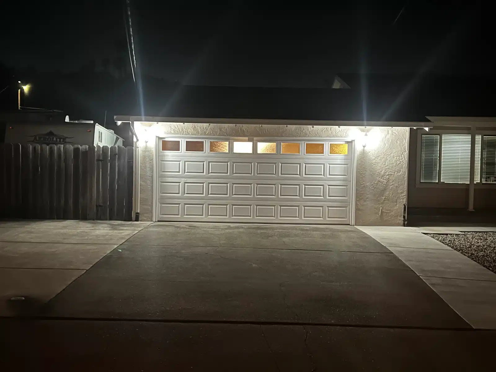 A white traditional raised-panel garage door with illuminated top windows glows under exterior lights at night, emphasizing the security provided by professional garage door repair.